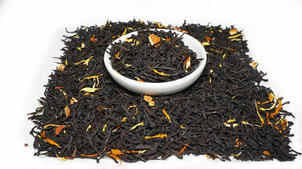 Black tea loose leaves with scattered orange-yellow citrus peel pieces, arranged on a white background with a small white bowl in the center filled with the tea.