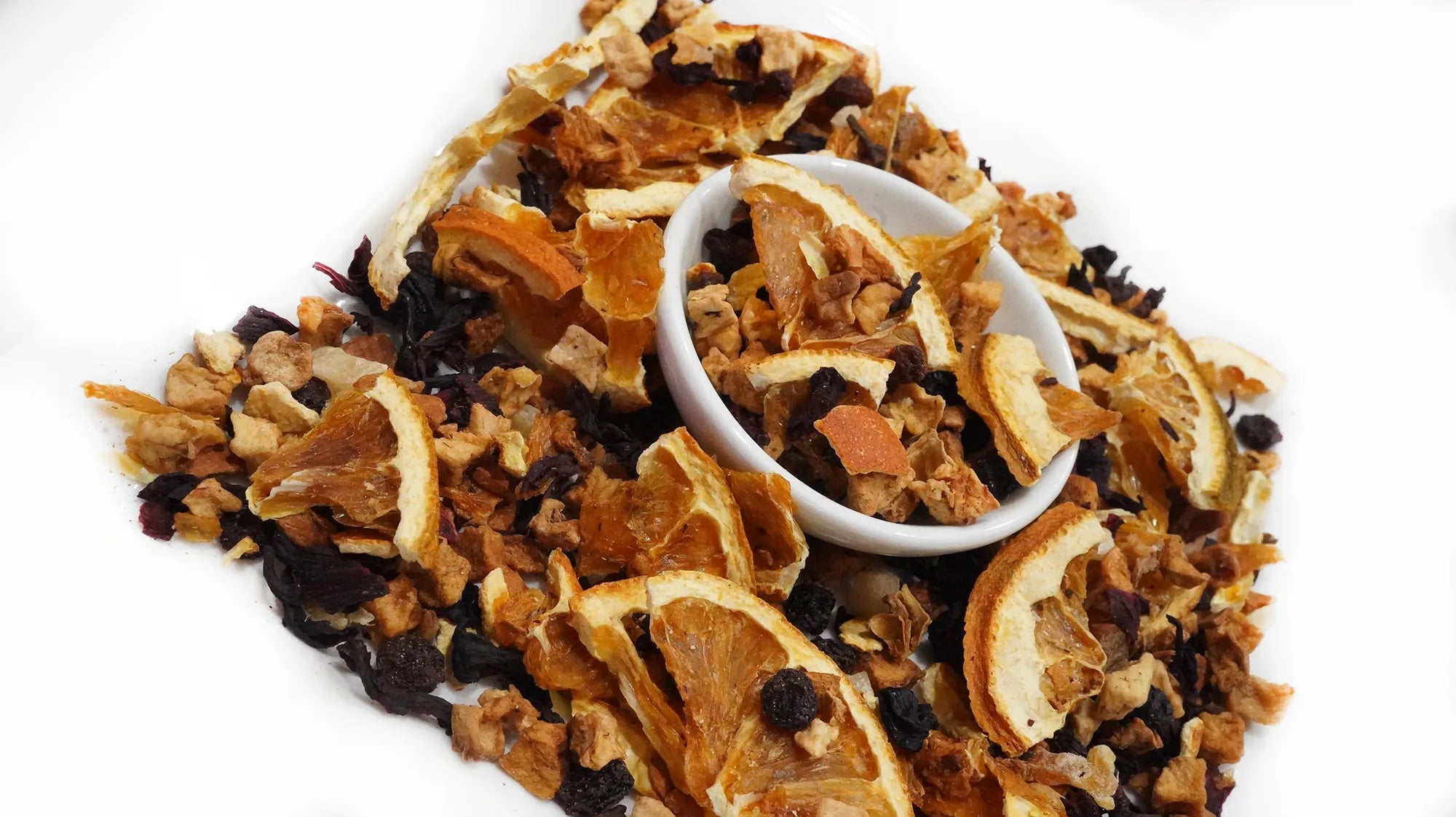 A pile of dried citrus and berry tea mix with a small white bowl in the center, photographed on a plain white background.
