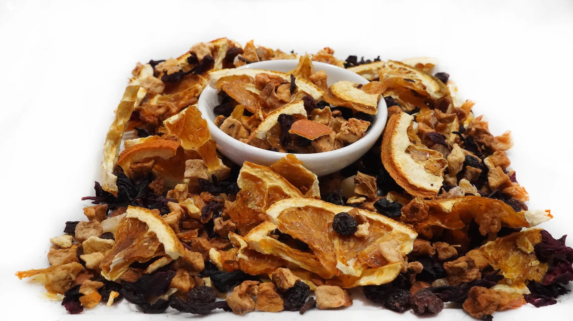 A pile of dried fruit and nut tea mix with a small white bowl in the center, photographed on a clean white background.