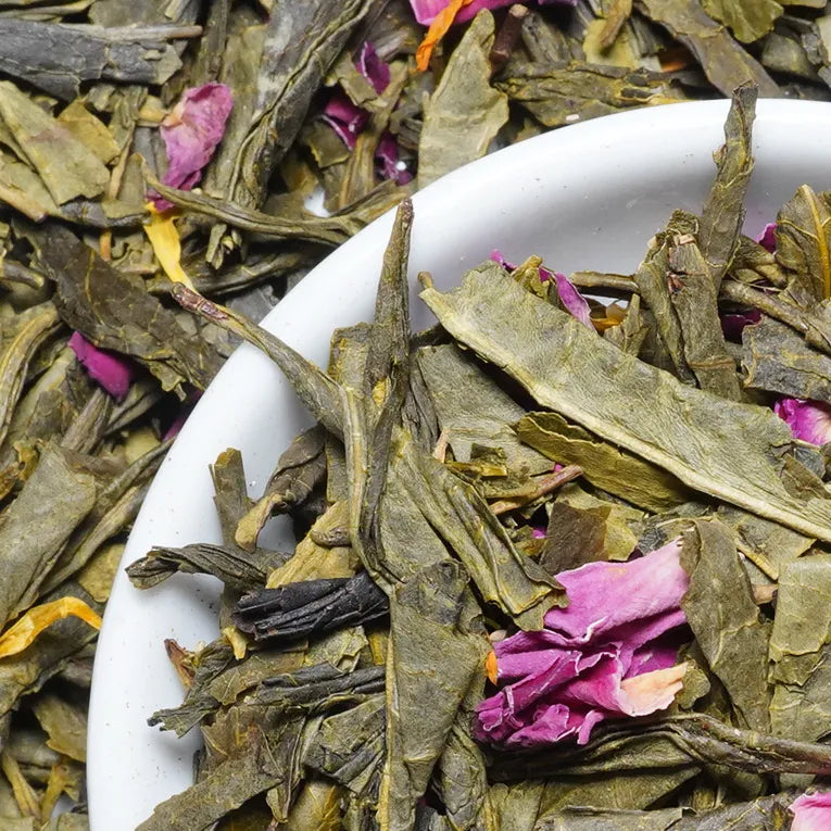 Close-up of loose dried tea leaves mixed with pink rose petals inside a white bowl, with more tea leaves filling the background.