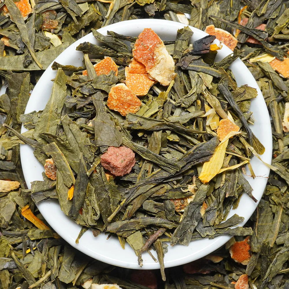 A top-down view of loose-leaf herbal tea inside a small white ceramic bowl, surrounded by more of the same tea leaves with dried orange and pink fruit pieces.