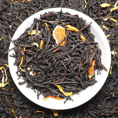 Dark loose-leaf tea blend in a white round dish, with visible dried citrus peel pieces and small yellow botanical accents, photographed from above against a matching tea-filled background.

