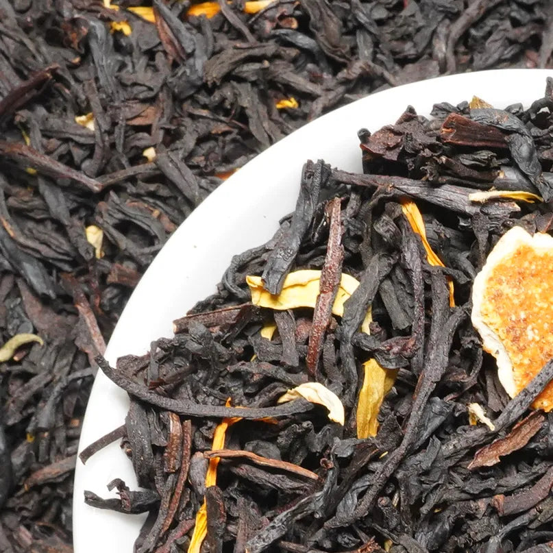 Close-up of loose-leaf tea in a white dish, showing dark brown to black tea leaves with small yellow dried flower or citrus slices.

