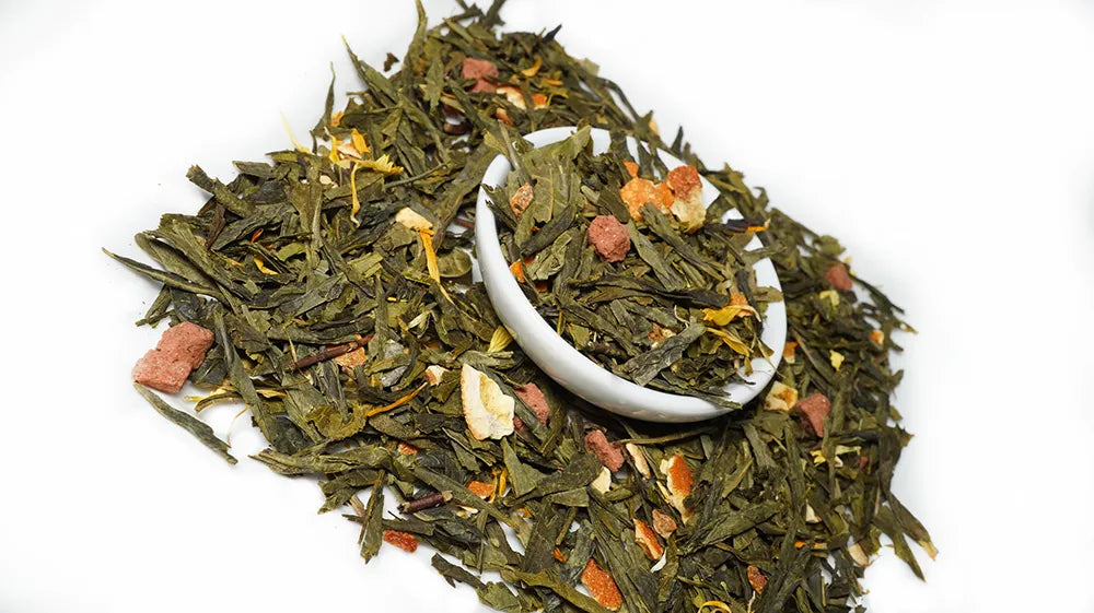 A pile of loose-leaf herbal tea with dried green leaves, orange flower petals, and small pinkish-brown pieces, with a small white scoop resting in the center, on a plain white background.

