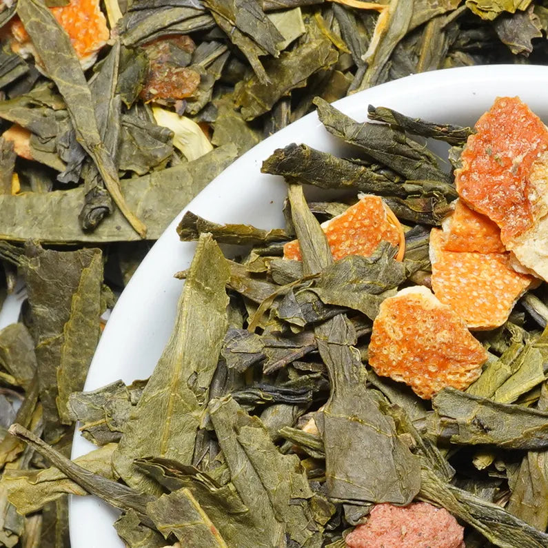 Close-up of loose-leaf tea with dark green leaves and bright orange dried fruit pieces, with some tea piled in a white bowl.


