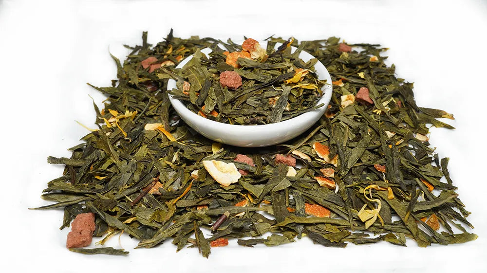 A pile of loose-leaf herbal tea with dried green leaves, orange slices, and small pinkish pieces, with a small white bowl placed in the center on a white background.

